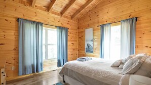 A wooden-paneled room with a bed, two windows with curtains, and a small rug. - Scenic Sunset Cabin RRG | Central | Hot Tub | Pets (Campton)