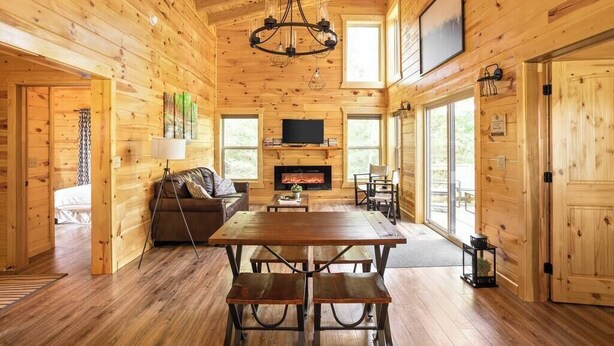 A wooden cabin interior with a dining table, benches, a sofa, a fireplace, and a television. - Scenic Sunset Cabin RRG | Central | Hot Tub | Pets (Campton)