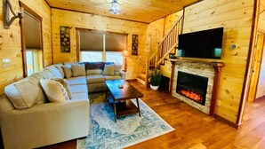 A cozy log cabin interior with a sectional sofa, a fireplace, a TV mounted above it, and a staircase leading to an upper level.