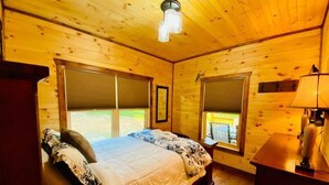 A wooden cabin bedroom with a bed, two windows, a desk, and a lamp.