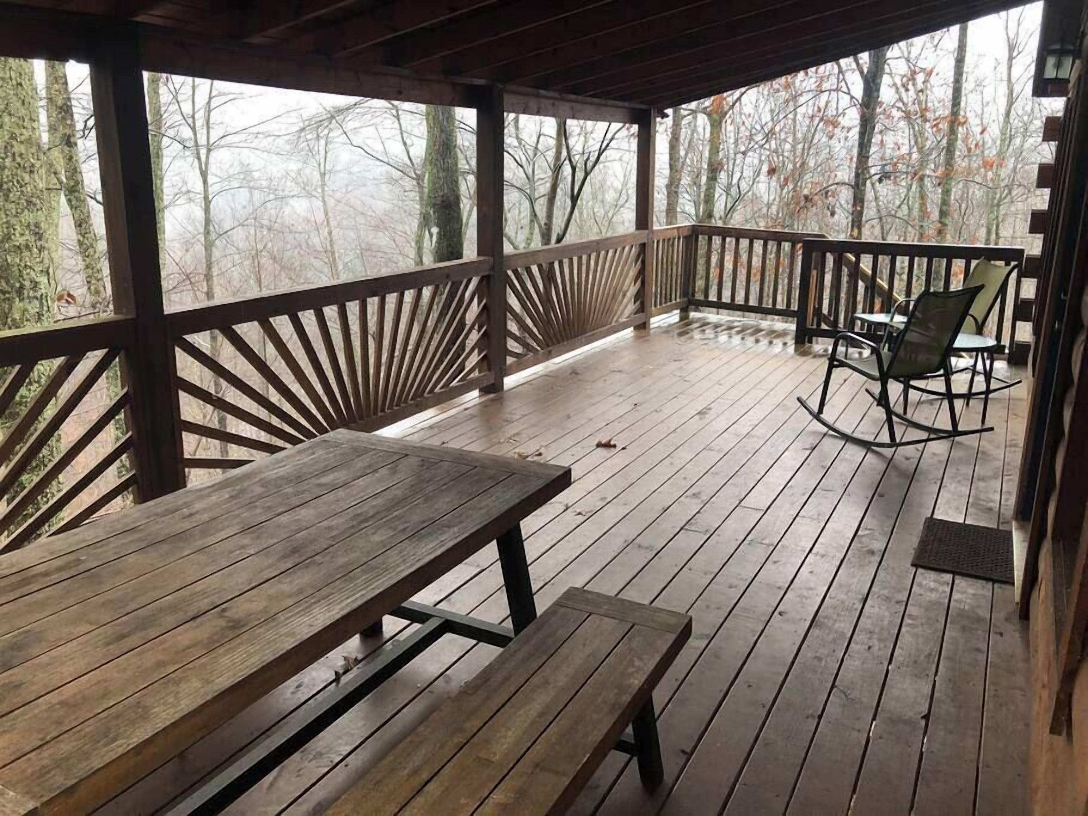 Een houten terras met een picknicktafel en een schommelstoel, omringd door bomen.