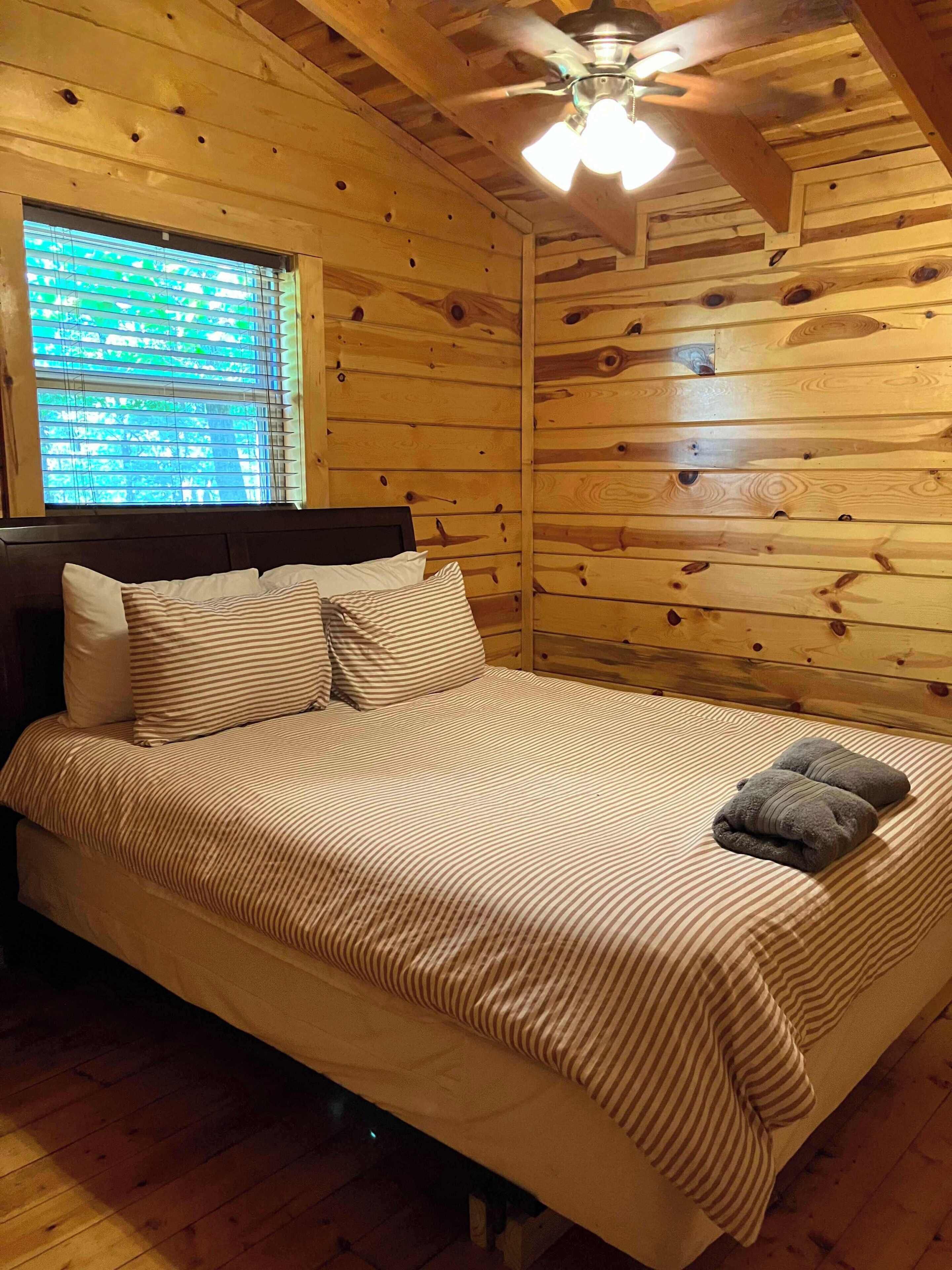 A wooden cabin room with a bed, a ceiling fan, and a window with blinds.