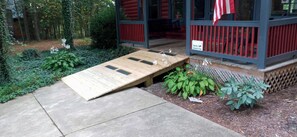 A wooden ramp leading to a porch with a red railing, surrounded by greenery and a small sign.