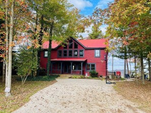 A red cabin with a porch, surrounded by trees and a gravel driveway.