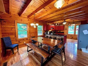 A wooden cabin interior with a dining table, benches, and a kitchen area.