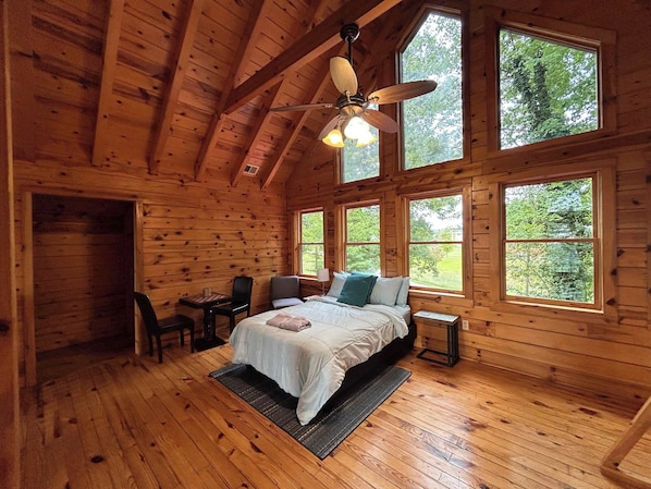 A wooden cabin bedroom with a bed, a chair, a desk, and a ceiling fan.