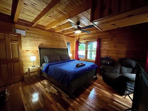 Camera da letto in una baita di legno con un letto, una sedia, un comodino e una ventola a soffitto.