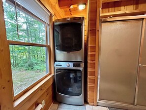 Una compatta cabina in legno con un'ampia finestra che si affaccia su una foresta, una lavatrice e asciugatrice affiancate, e una porta con zanzariera.