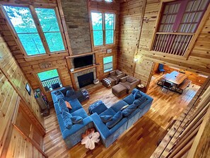 A spacious cabin interior with a large sofa, a fireplace, and multiple windows offering views of the surrounding forest.