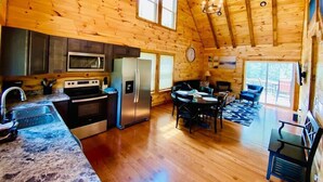 A wooden cabin interior with a kitchen, dining area, and a large window.