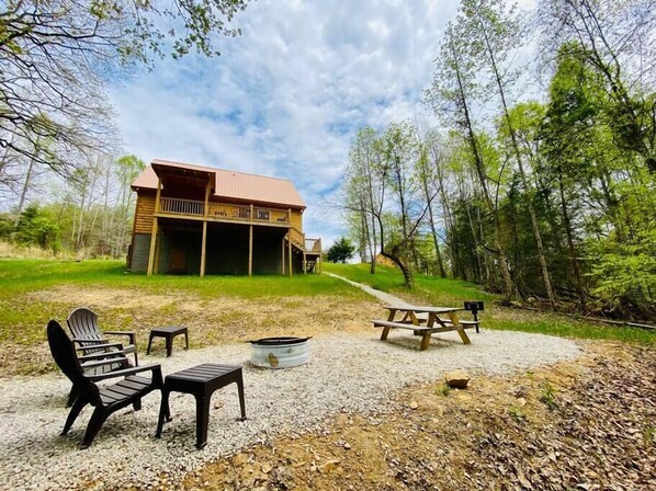 A rustic cabin with a porch, surrounded by trees and a gravel area with picnic tables and chairs. - Timberview - Central RRG with WiFi, Hot Tub, Fire Pit (Pine Ridge)