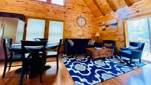 A wooden cabin interior with a dining table, chairs, a sofa, a clock, and a large window.