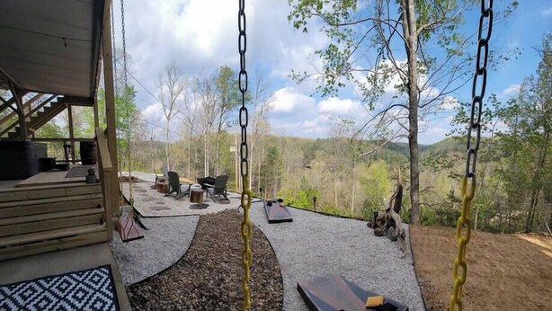 A wooden deck with outdoor seating, a swing set, and a view of a forested landscape. - Walden’s Wonder – Artful Cabin w/ Valley View (Pine Ridge, KY)
