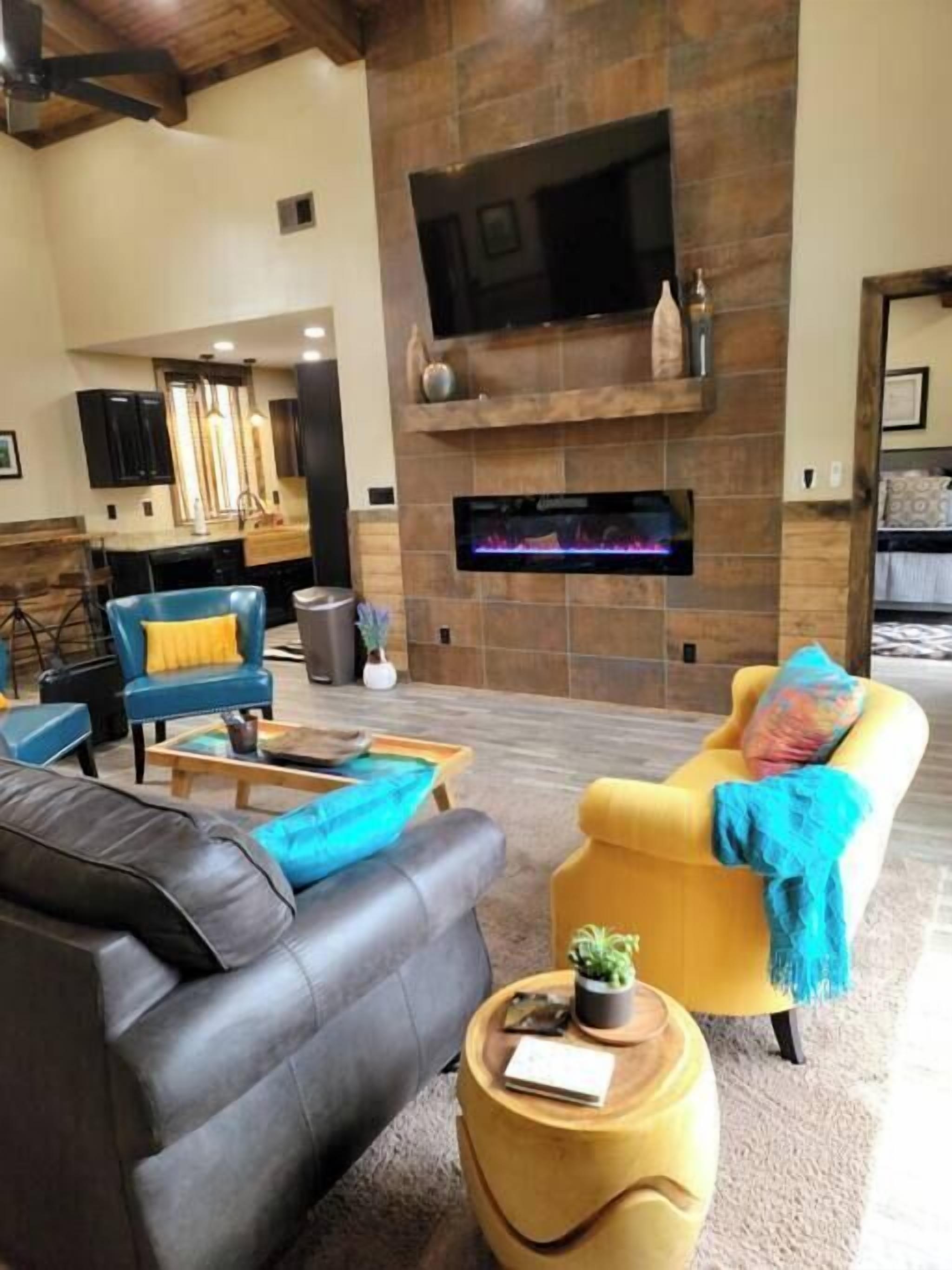 A living room with a fireplace, a TV mounted above it, and a kitchen area in the background.