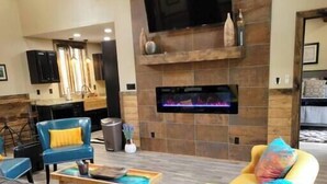 A living room with a fireplace, a TV mounted above it, and a kitchen area in the background. - Walden’s Wonder – Artful Cabin w/ Valley View (Pine Ridge, KY)