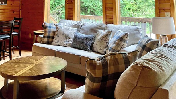 A cozy cabin interior with a wooden ceiling, a sofa with plaid cushions, a round wooden table, and large windows offering a view of the forest. - Wildcat Cabin • Pet Friendly, Hot Tub, Forest Stay (Stanton)