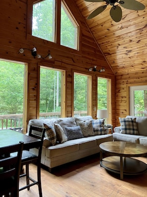 Cozy cabin with a high ceiling, exposed beams, and large windows offering a forest view. Features a sofa set, dining table, and coffee table.