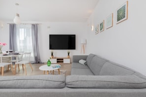 A modern living room with a grey sofa, a flat-screen TV, a dining area with a white table and chairs, and a small coffee table.