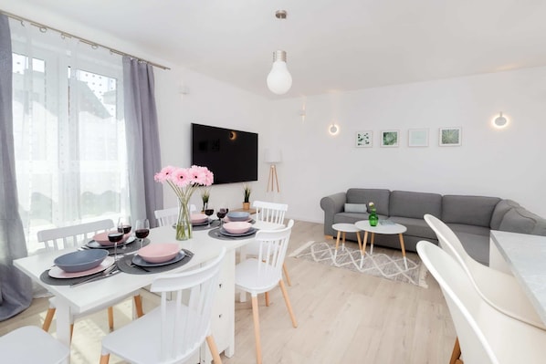 A modern living room with a grey sofa, a dining table set with pink and grey plates, and a flat-screen TV mounted on the wall.