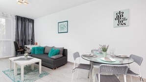 Un salon moderne avec un canapé gris, une table basse blanche et une table à manger en verre avec des chaises. Un tableau encadré orne le mur, et une fenêtre est agrémentée de rideaux.