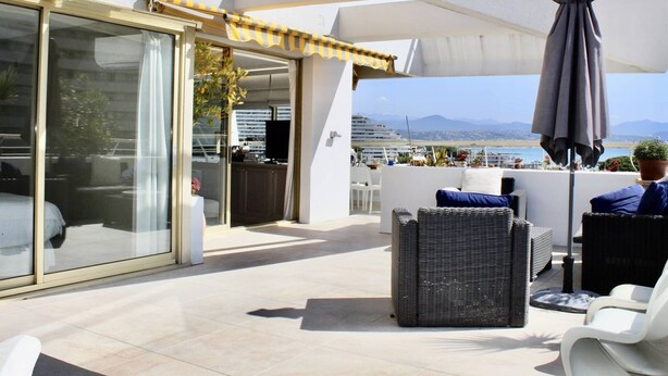 A rooftop terrace with a view of the sea and mountains, featuring outdoor seating, a large umbrella, and a glass door leading to an interior space.