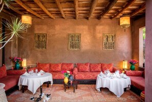 A cozy room with a wooden ceiling, two sofas with red cushions, a round table with a white tablecloth, and decorative wall art. - Jnane Leila (Marrakech)