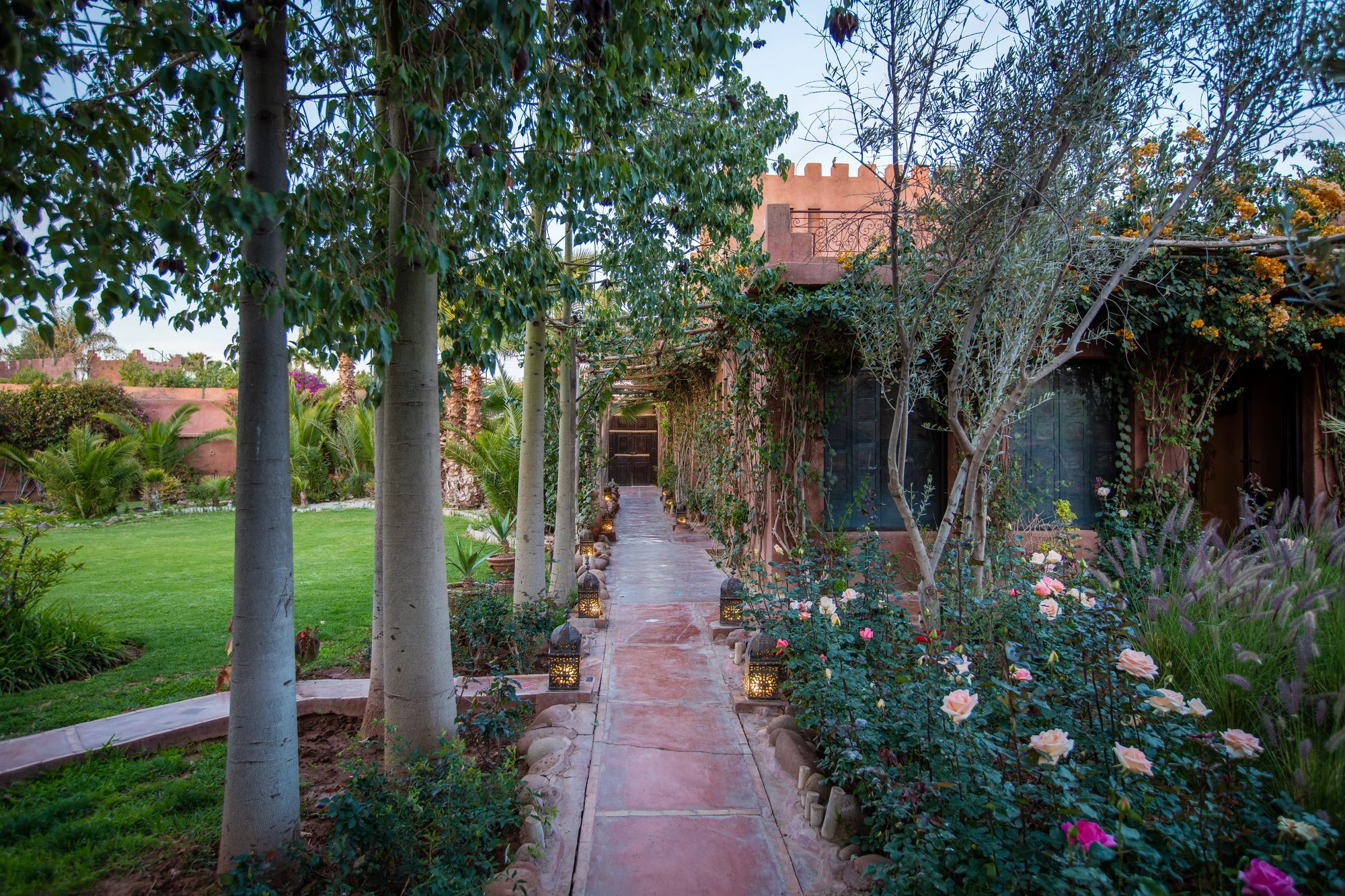 Un sendero en un jardín que conduce a un edificio de estilo arquitectónico tradicional, rodeado de exuberante vegetación y plantas con flores.
