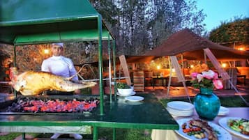 Un chef asando un pescado grande en una parrilla al aire libre, con mesas y sillas dispuestas debajo de una carpa.