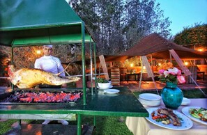 A chef grilling a large fish on an outdoor grill, with dining tables and chairs set up under a tent. - Jnane Leila (Marrakech)