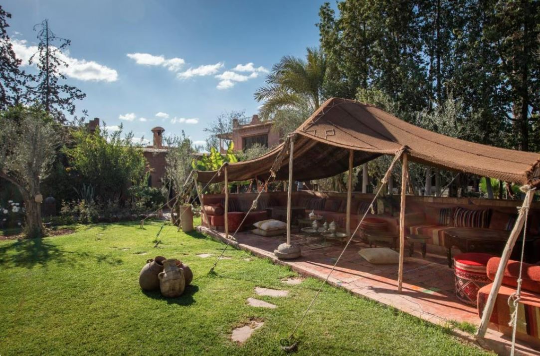 Un área de estar al aire libre rústica con un toldo, rodeada de vegetación y un edificio de ladrillo al fondo.