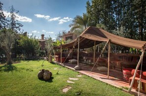 A rustic outdoor seating area with a canopy, surrounded by greenery and a brick building in the background. - Jnane Leila (Marrakech)