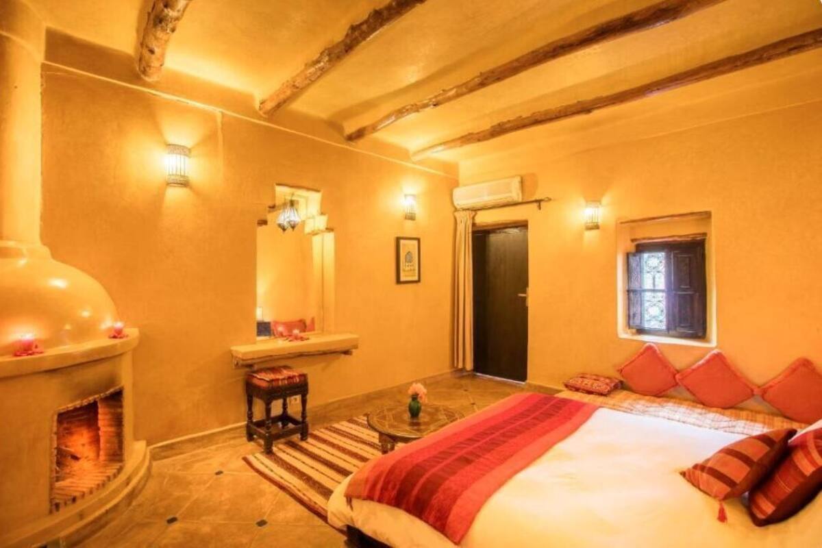 A cozy bedroom with a fireplace, a bed with red bedding, and a small table with a vase.