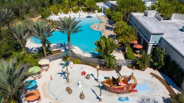 Luftaufnahme eines Resorts mit einem zentralen Schwimmbecken, umgeben von Palmen und Liegestühlen.