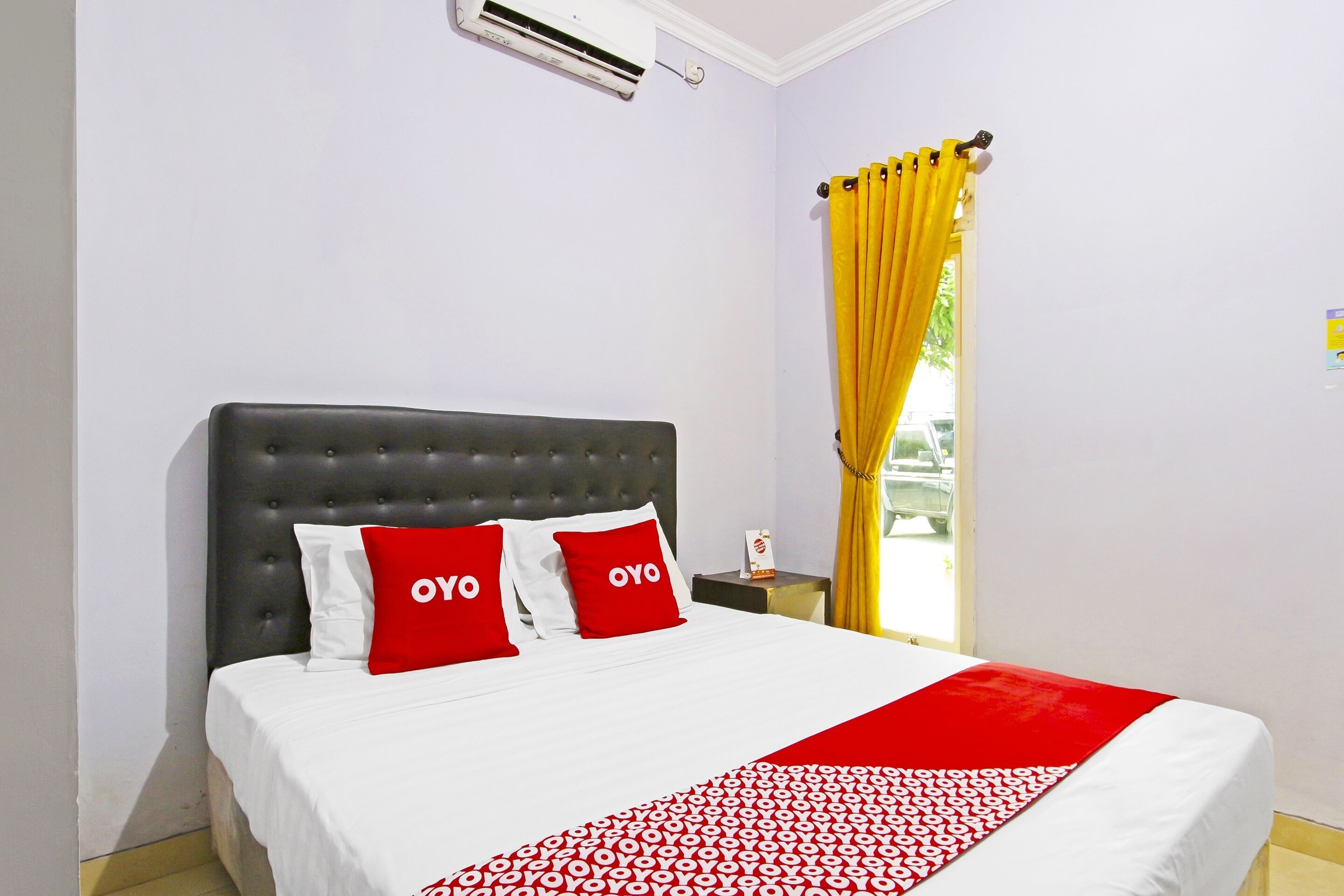 Quarto de hotel com uma cama, cabeceira, mesa de cabeceira, janela com cortinas amarelas e um ar condicionado na parede.