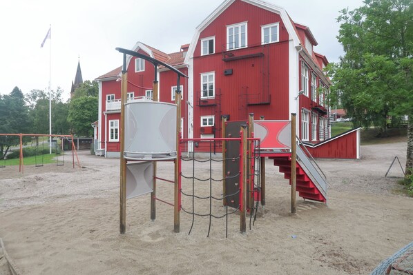 A playground with a slide and climbing structures, a red building in the background, and a flagpole. - SKRÅ hostel- business bed & breakfast (Alno)