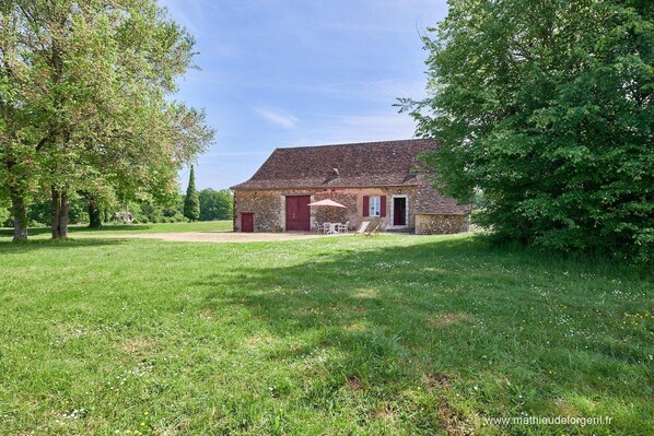 Un bâtiment rustique en pierre, avec une porte rouge, entouré d’une pelouse verdoyante et d’arbres.