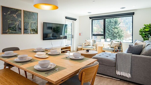 A modern living room with a dining area, large windows, and a grey sofa.
