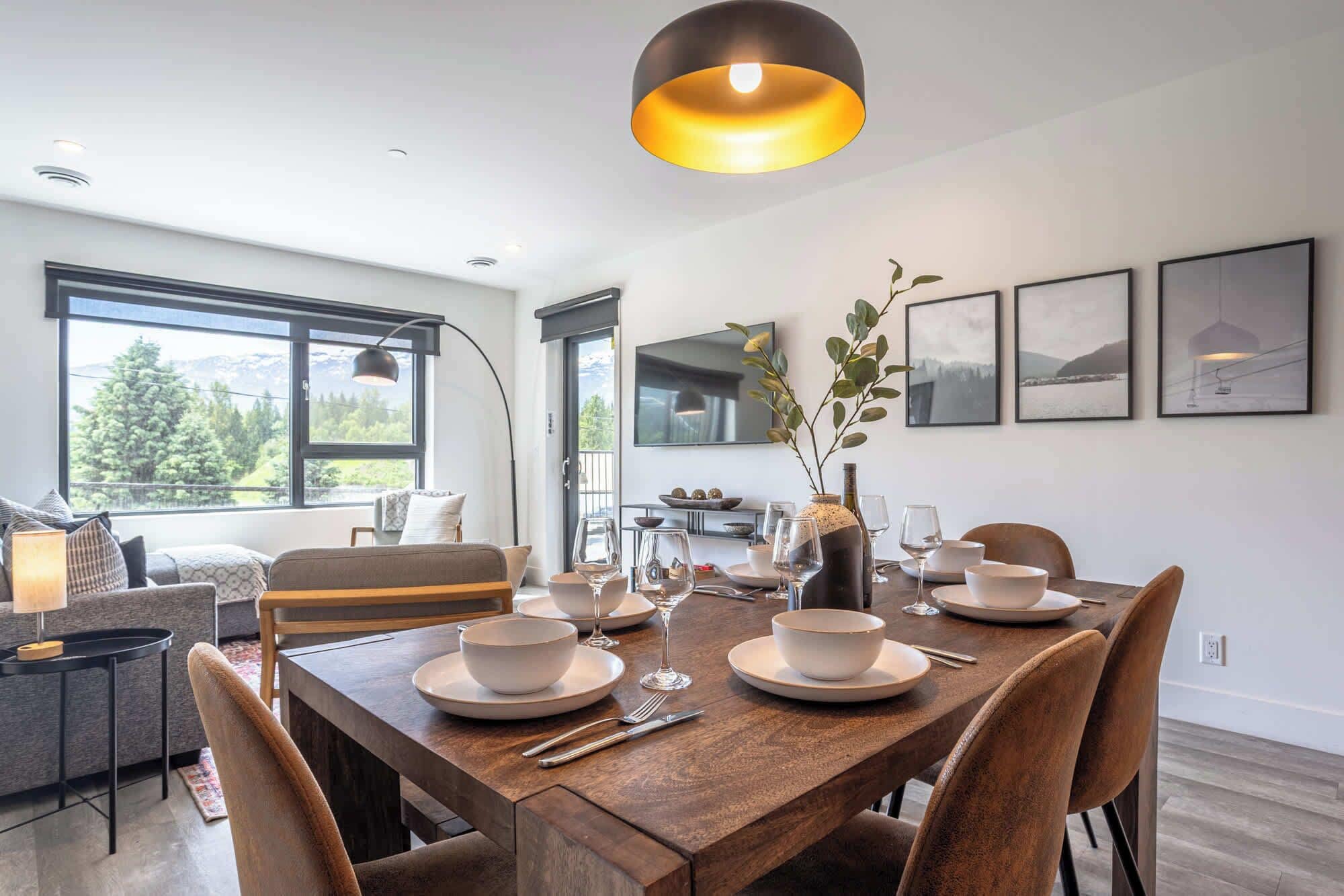 Un comedor con una mesa de madera dispuesta para cuatro personas, un sofá, un televisor y cuadros en la pared.