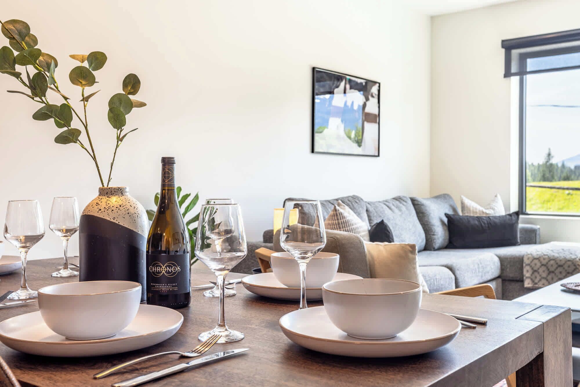 Comedor con una mesa dispuesta para cuatro personas, una botella de vino y un florero con hojas verdes.
