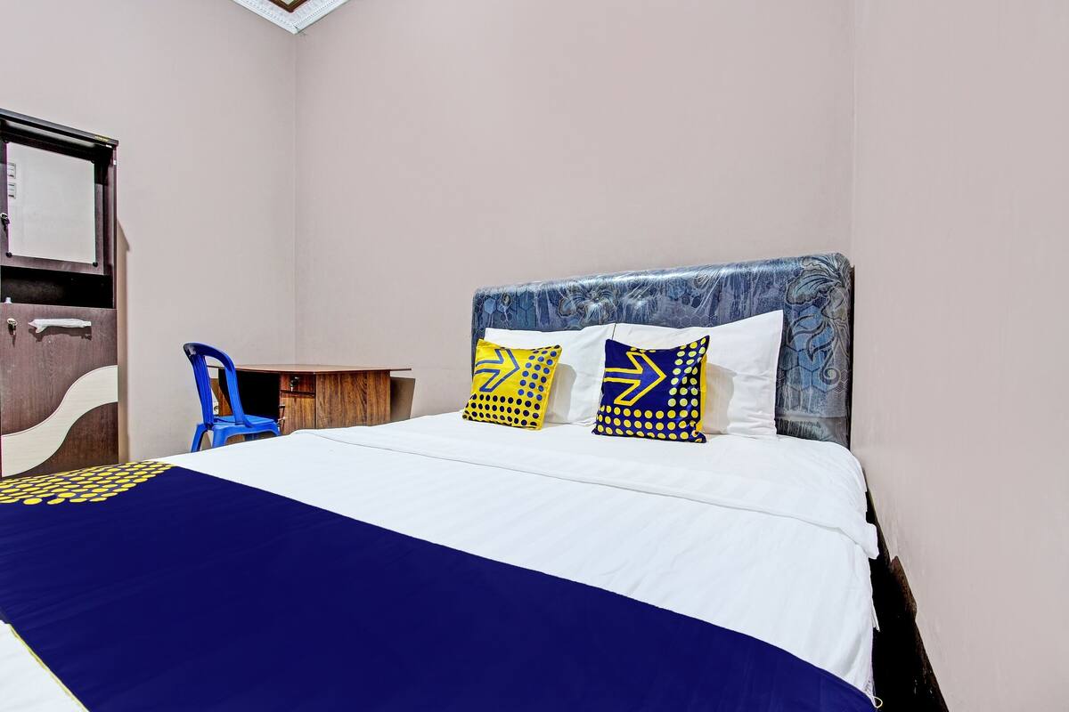 Ein ordentlich bezogenes Bett mit einer blauen und weißen Tagesdecke, zwei Zierkissen mit geometrischen Mustern, ein blauer Sessel und ein Nachttisch aus Holz.