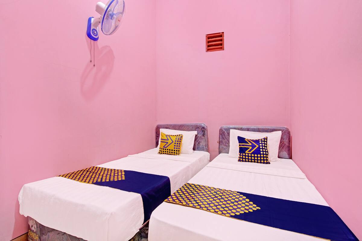 Zwei Einzelbetten mit weißer und blauer Bettwäsche, ein kleiner Ventilator und ein roter Lüftungsgitter an einer rosafarbenen Wand.