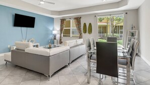A modern living room with a sofa, a glass coffee table, a dining table with chairs, a flat-screen TV mounted on the wall, and a ceiling fan. - 9817 Cristalino Way #102 (Fort Myers)