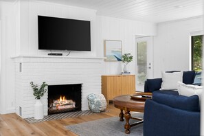 A living room with a white brick fireplace, a flat-screen TV mounted above it, a blue sofa, and a wooden cabinet. - Three Pines Hideaway (Glen Arbor)