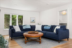 Una sala de estar con un sofá seccional azul oscuro, una mesa de centro redonda de madera y un ventanal con vistas a árboles.