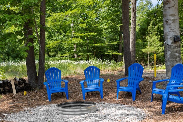 A forested area with three blue Adirondack chairs arranged around a fire pit. - Three Pines Hideaway (Glen Arbor)
