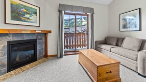 A living room with a fireplace, a sofa, a wooden chest, and a large window with a view of a building and trees.