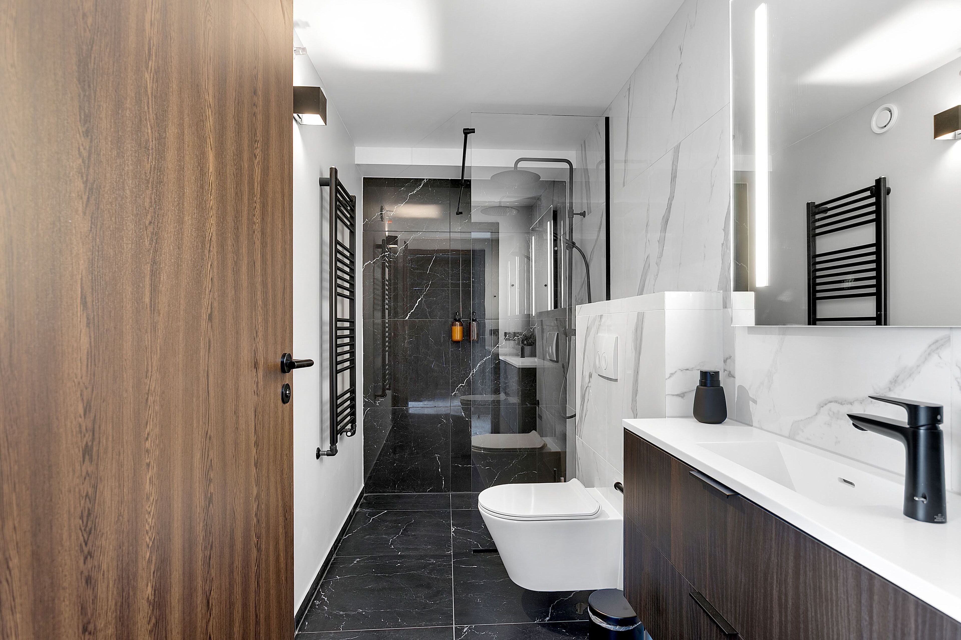 A modern bathroom with a wooden door, a white toilet, a black bathtub, and a marble countertop.