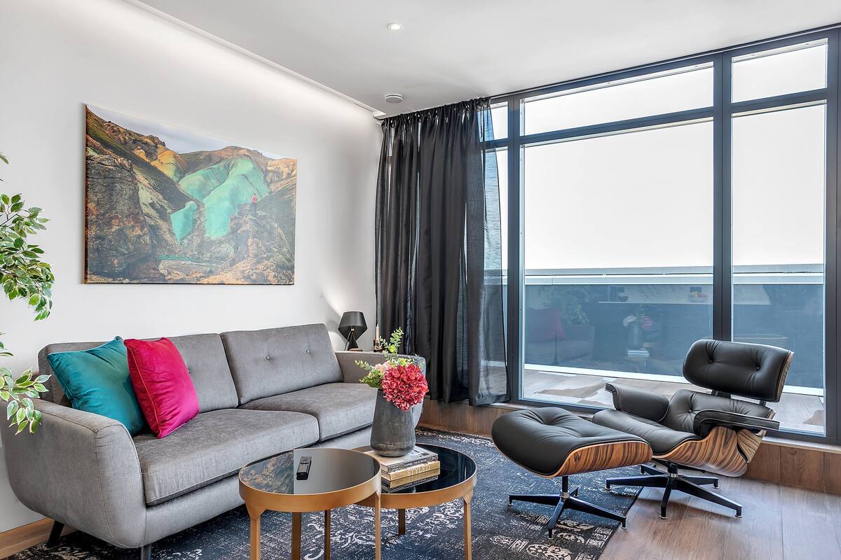 Een moderne woonkamer met een grijze bank, een zwarte lederen fauteuil, een glazen salontafel en een groot raam met gordijnen.