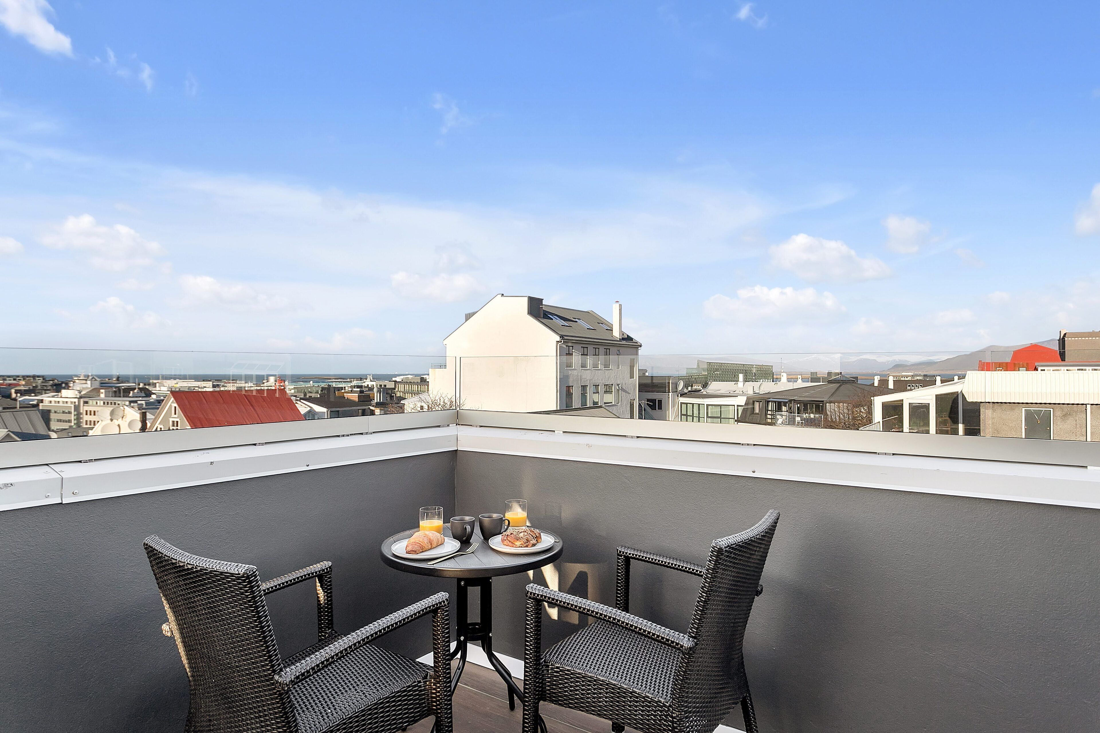 Un balcon avec des fauteuils en osier et une table, d’où l’on peut admirer un panorama urbain avec des bâtiments et un ciel dégagé.