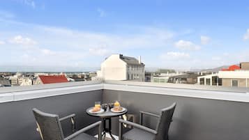A balcony with wicker chairs and a table, overlooking a cityscape with buildings and a clear sky.